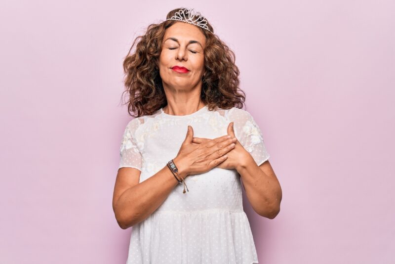 Vrouw van middelbare leeftijd met kroontje (queenager) legt haar beide handen op haar hart. Een symbool van zelfacceptatie.